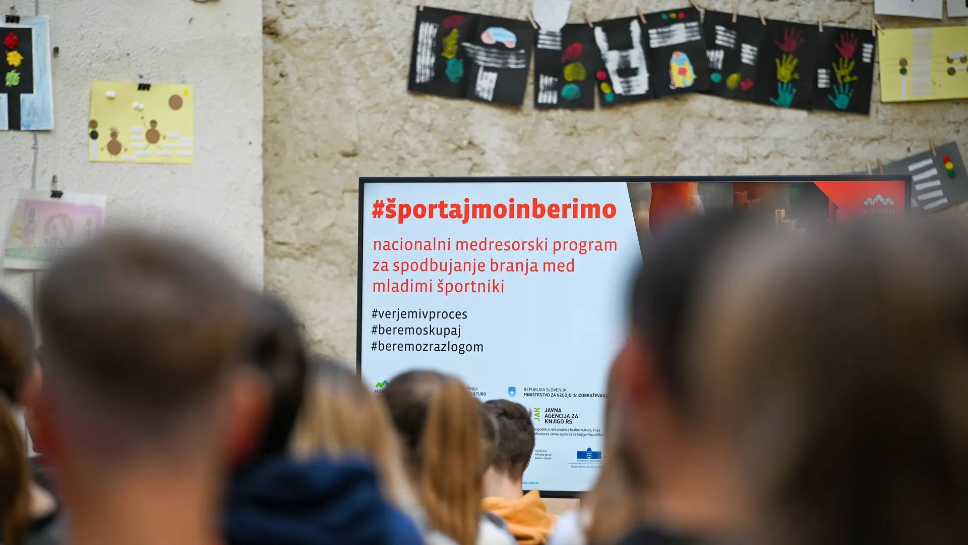 Dijaki sedijo v prostoru in gledajo zaslon s predstavitvijo programa #športajmoinberimo, nacionalnega medresorskega programa za spodbujanje branja med mladimi športniki; v ozadju so na steni razstavljeni likovni izdelki.