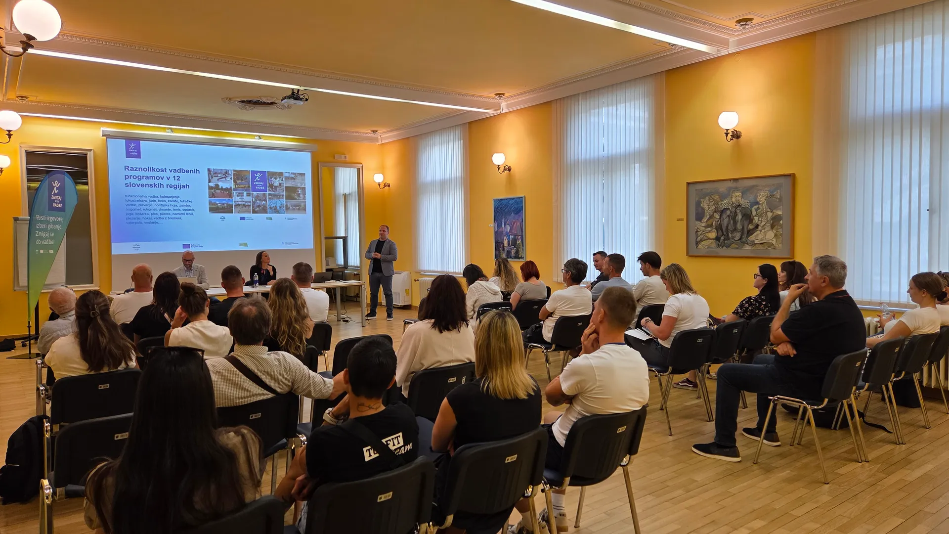 Raznolikost vodenih programov v 12 slovenskih regijah je predstavljena na projekciji, medtem ko poslušalci sedijo v stolih.