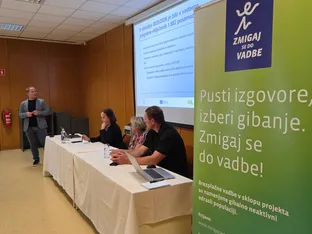 Na odru so štirje ljudje, eden govori, ostali pa sedijo za mizo z računalniki in steklenicami vode, ob strani je plakat z informacijami.