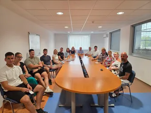 V prostoru je skupina ljudi, ki sedi ob dolgi mizi, nekateri imajo bele in temne obleke.