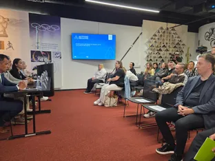Na odru sedi pet govorcev za mizo, v ozadju pa je skupina poslušalcev in projekcijski zaslon.