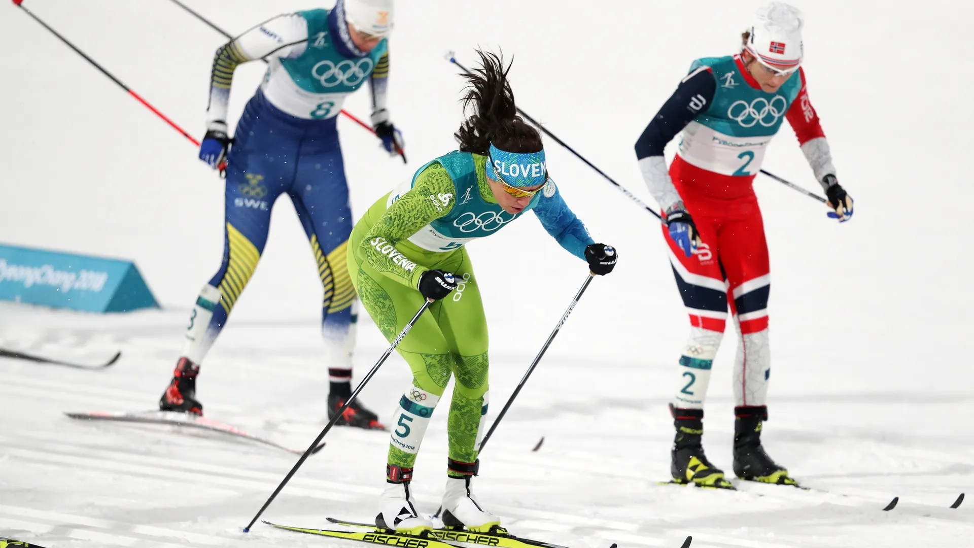 Tri smučarke na snežni progi, ena v zeleni opremi s številko 5, druga v modri in tretja v rdeči opremi.