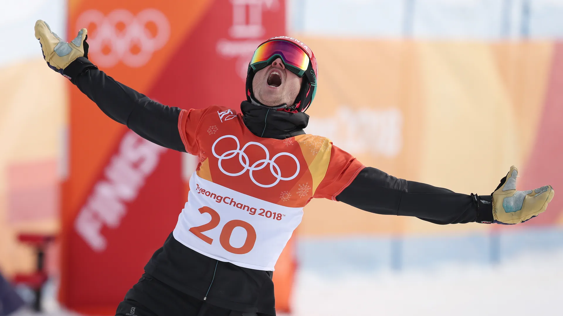 Športnik v rdeči majici s številko 20 in olimpijskimi krogi na prsih praznuje z razprtimi rokami in odprtimi usti.