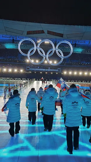 Na odru so udeleženci oblečeni v bele plašče, medtem ko se svetijo modri in zeleni vzorci ter olimpijski krogi visijo nad njimi.