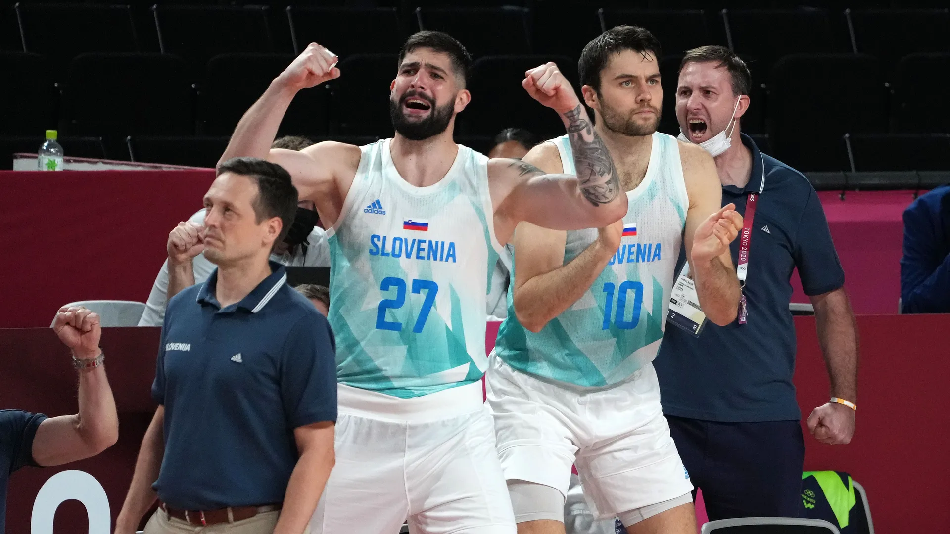 Dva košarkarja v belih dresih s številkama 27 in 10 ter več članov ekipe, ki izražajo navdušenje.