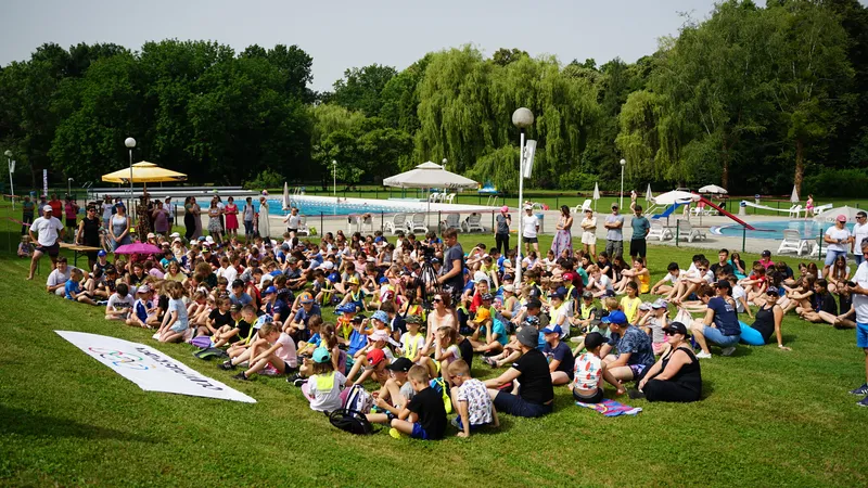 Za Olimpijski dan, na travnati površini sedi množica otrok in odraslih, ob robu pa je vidna transparent z napisom "Cilj je zmaga".
