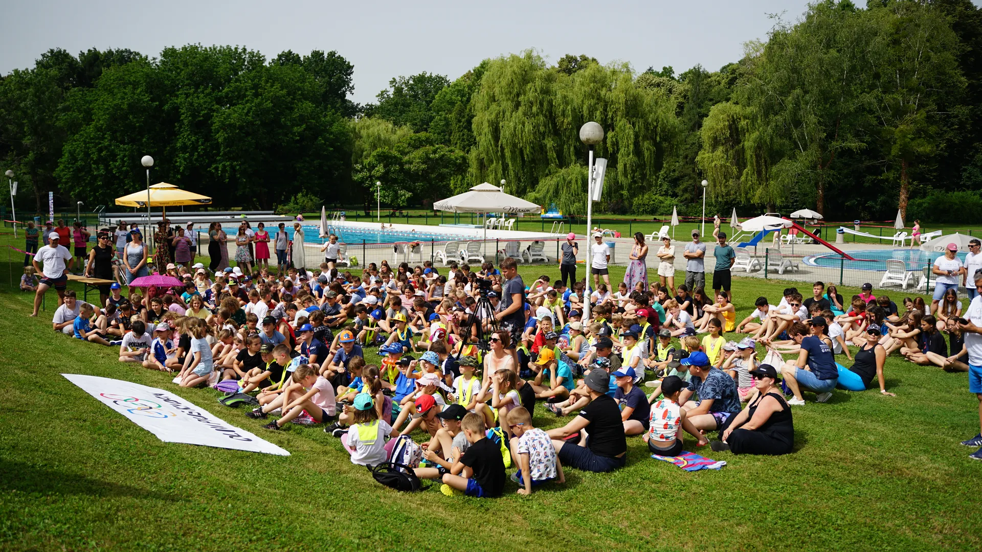 Za Olimpijski dan, na travnati površini sedi množica otrok in odraslih, ob robu pa je vidna transparent z napisom "Cilj je zmaga".
