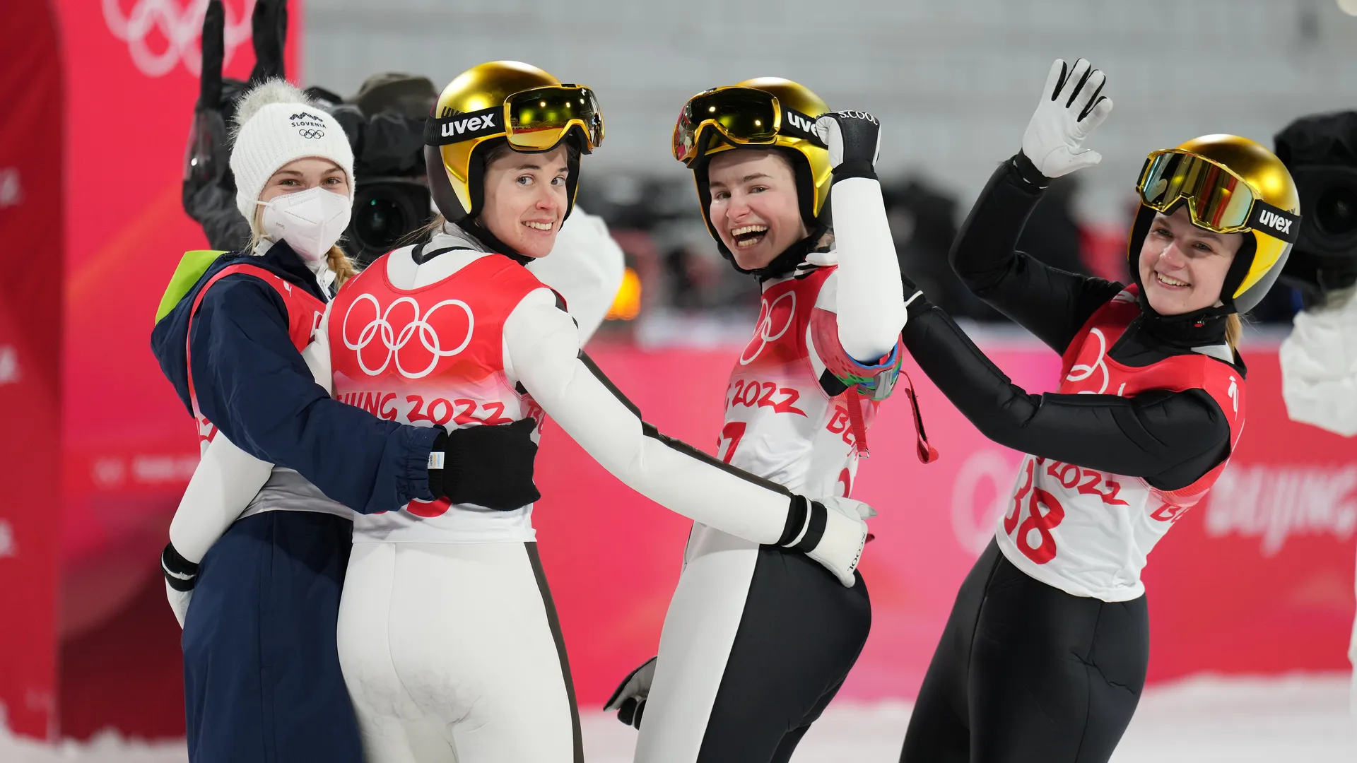 Štiri športnice v zimskih oblačilih z olimpijskimi čeladami in številkami na dresih se veselijo in pozdravljajo.
