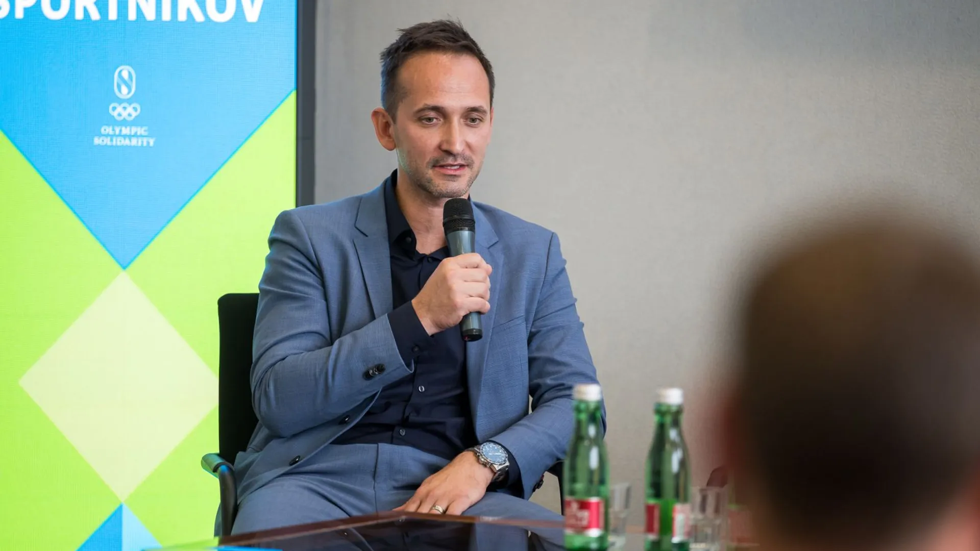 Moški v sivi obleki drži mikrofon in govori, medtem ko so na mizi steklenice vode.