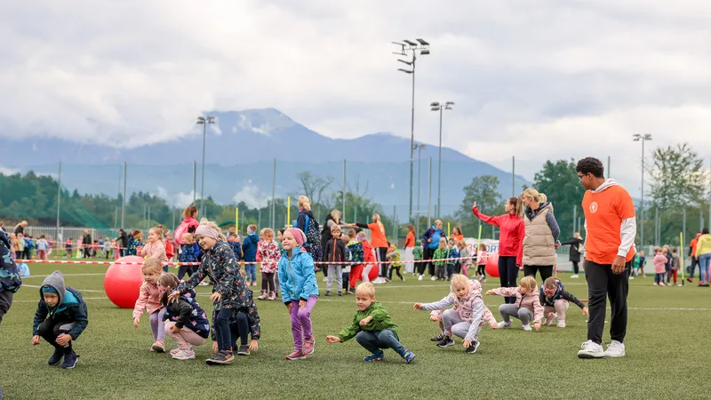 Otroci se igrajo na travnati površini, nekateri se sklanjajo, drugi stojijo, oblečeni so v različne barvite jakne.