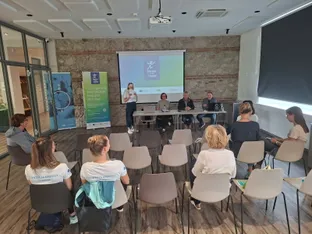 Na odru je govornica, trije sogovorniki sedijo za mizo, v ozadju so plakati in projekcija z besedilom "Zmigaj se do vadbe!.