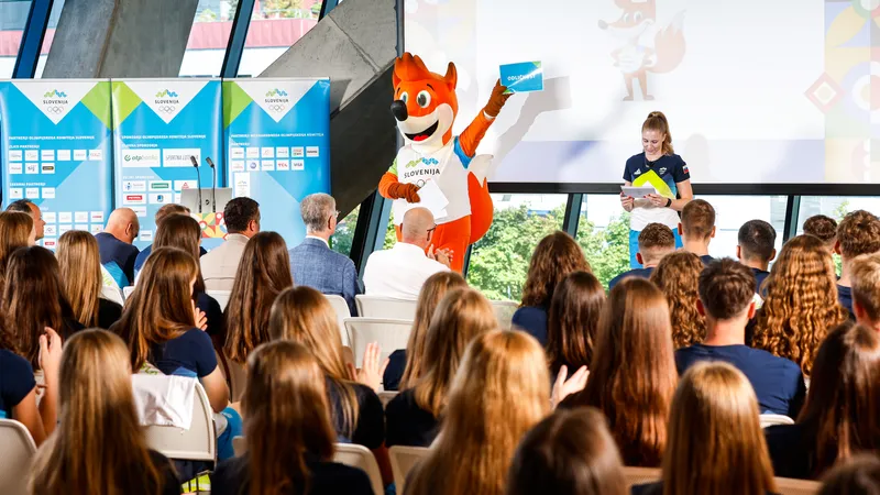 Maskota v oranžni obleki drži znak, medtem ko športnica v modri obleki bere papirje pred občinstvom.