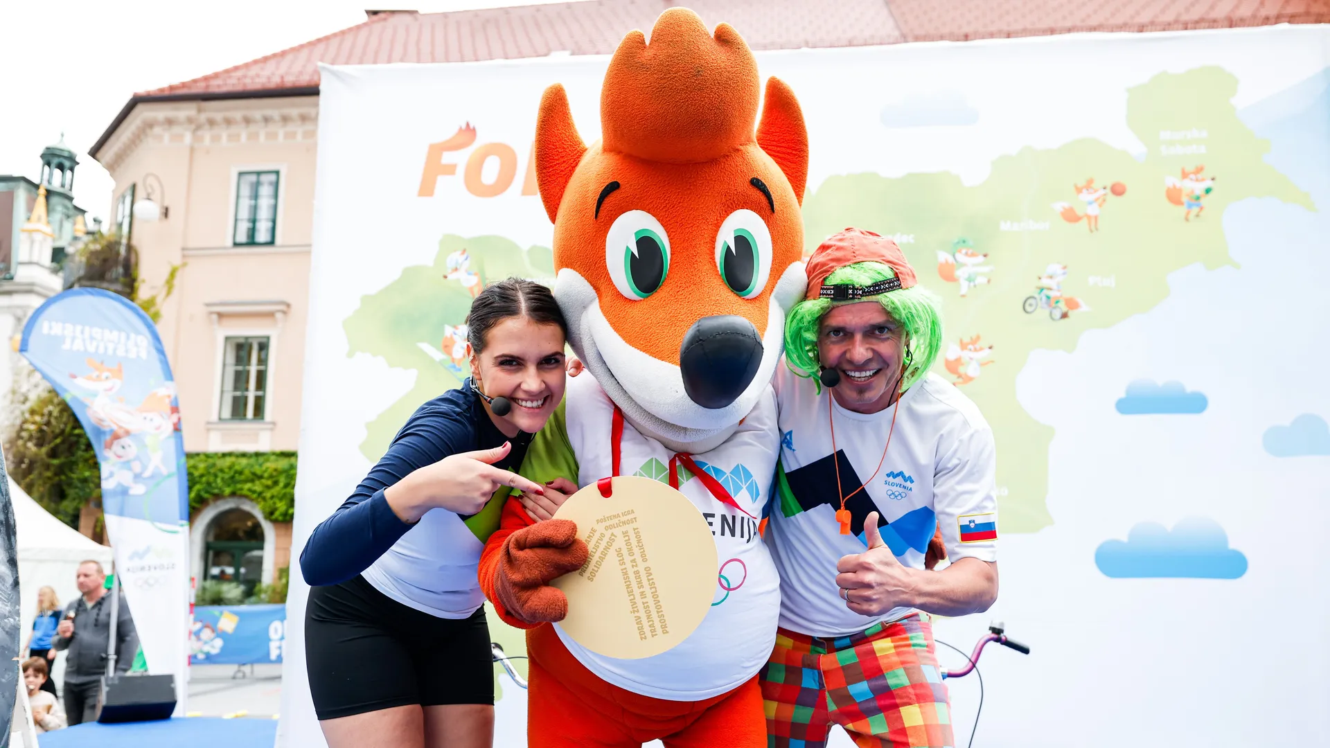 Ženska v športni obleki in moški v barvni obleki pozirata z maskoto Foksi, ki drži medaljo z napisom.
