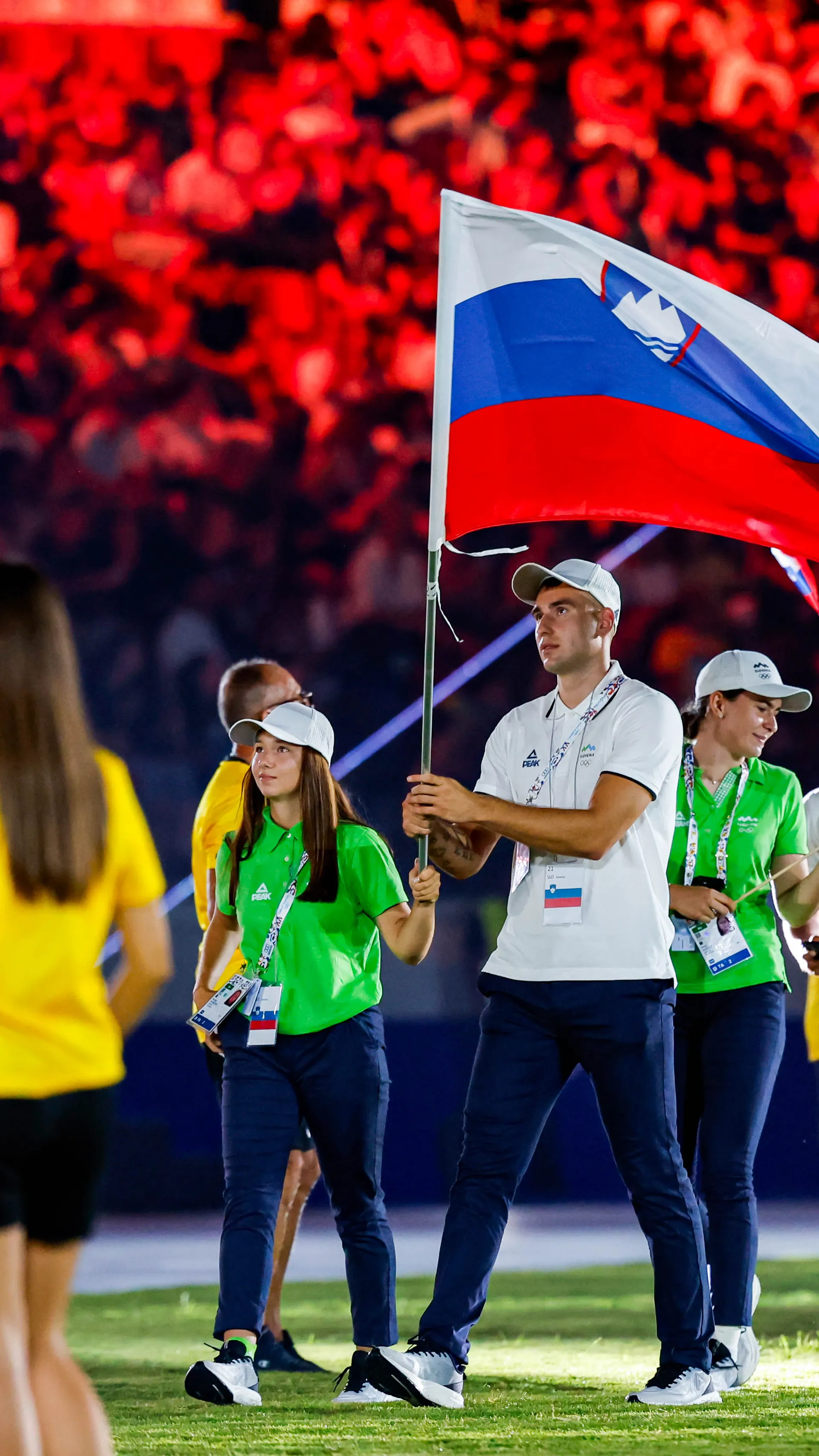 Moški drži zastavo Slovenije, obkrožen z mladimi športniki v belih in zelenih majicah.