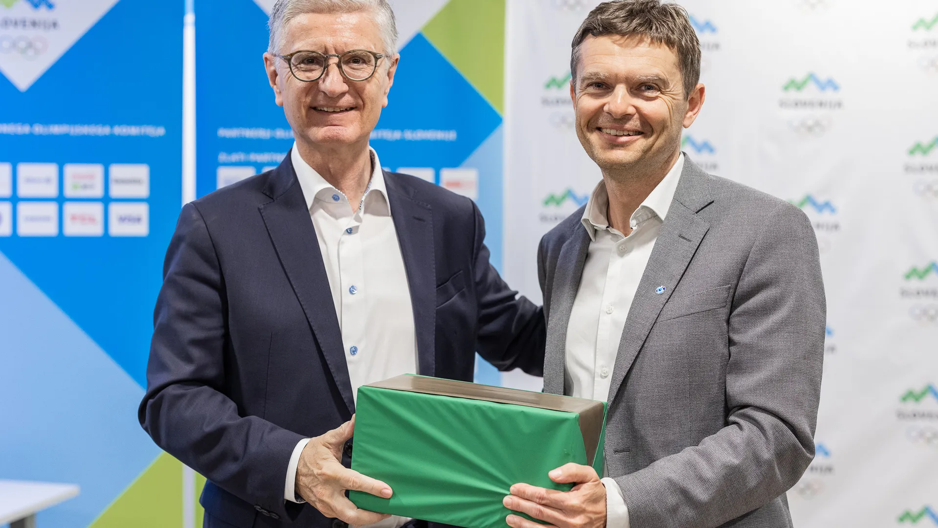 Dva moška, oblečena v poslovna oblačila, se smehljata in držita zeleno škatlo.