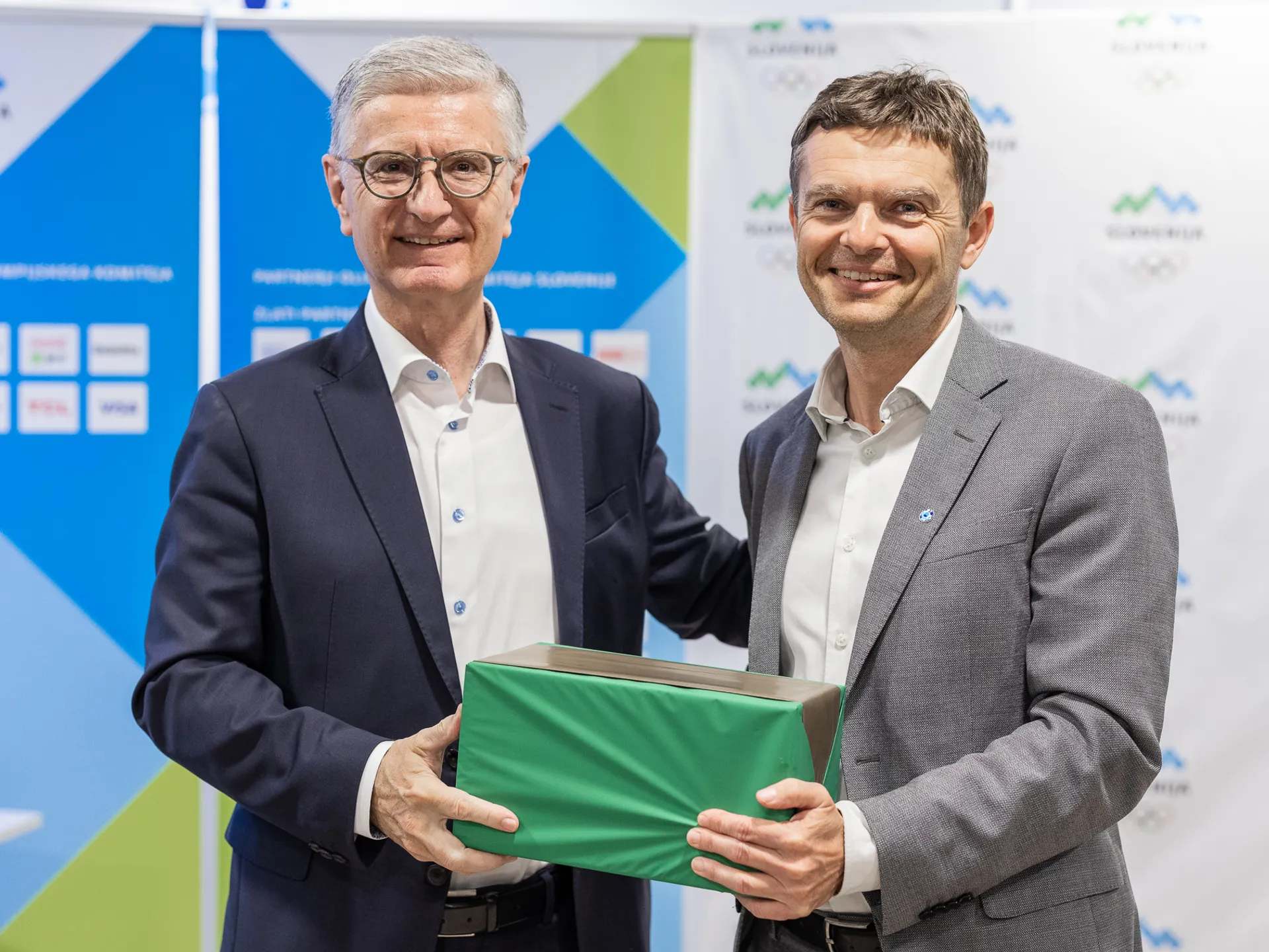 Dva moška, oblečena v poslovna oblačila, se smehljata in držita zeleno škatlo.