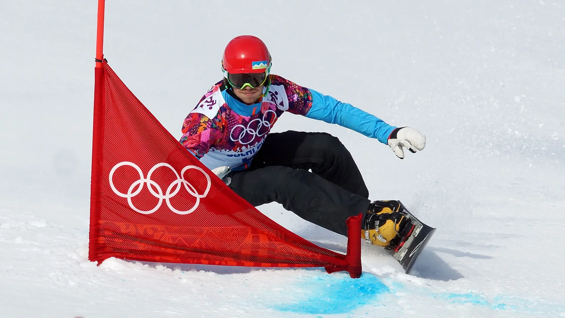 Žan Košir, slovenski smučar na snegu se nagiba ob rdeči olimpijski zastavi, oblečen v barvno majico in čelado.