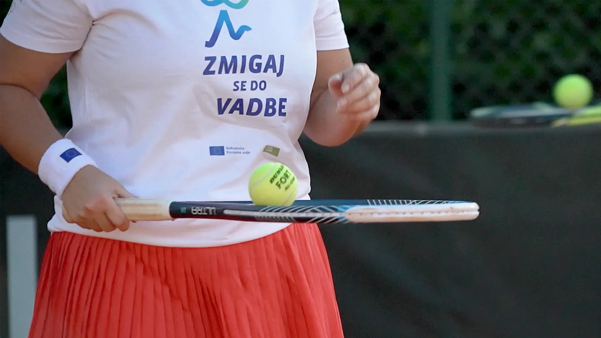 Ženska v beli majici z napisom "ZMIGAJ SE DO VADBE" drži teniško lopar in se pripravlja na udarec žoge.
