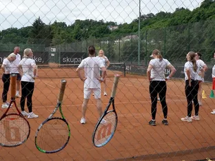 Na teniškem igrišču je skupina ljudi v belih majicah, ki se pogovarjajo in gledajo proti nebu.