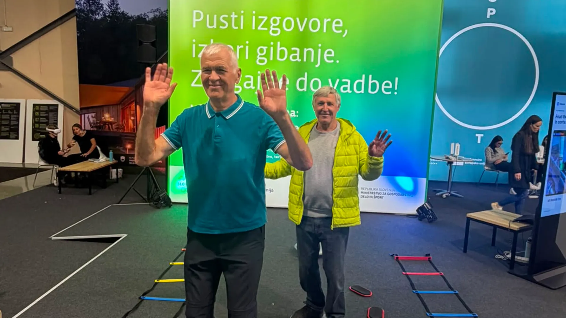 Dva moška stojita na ravnotežnih ploščah, eden nosi modro majico, drugi svetlo rumeno jakno, ob njiju so različni športni pripomočki.