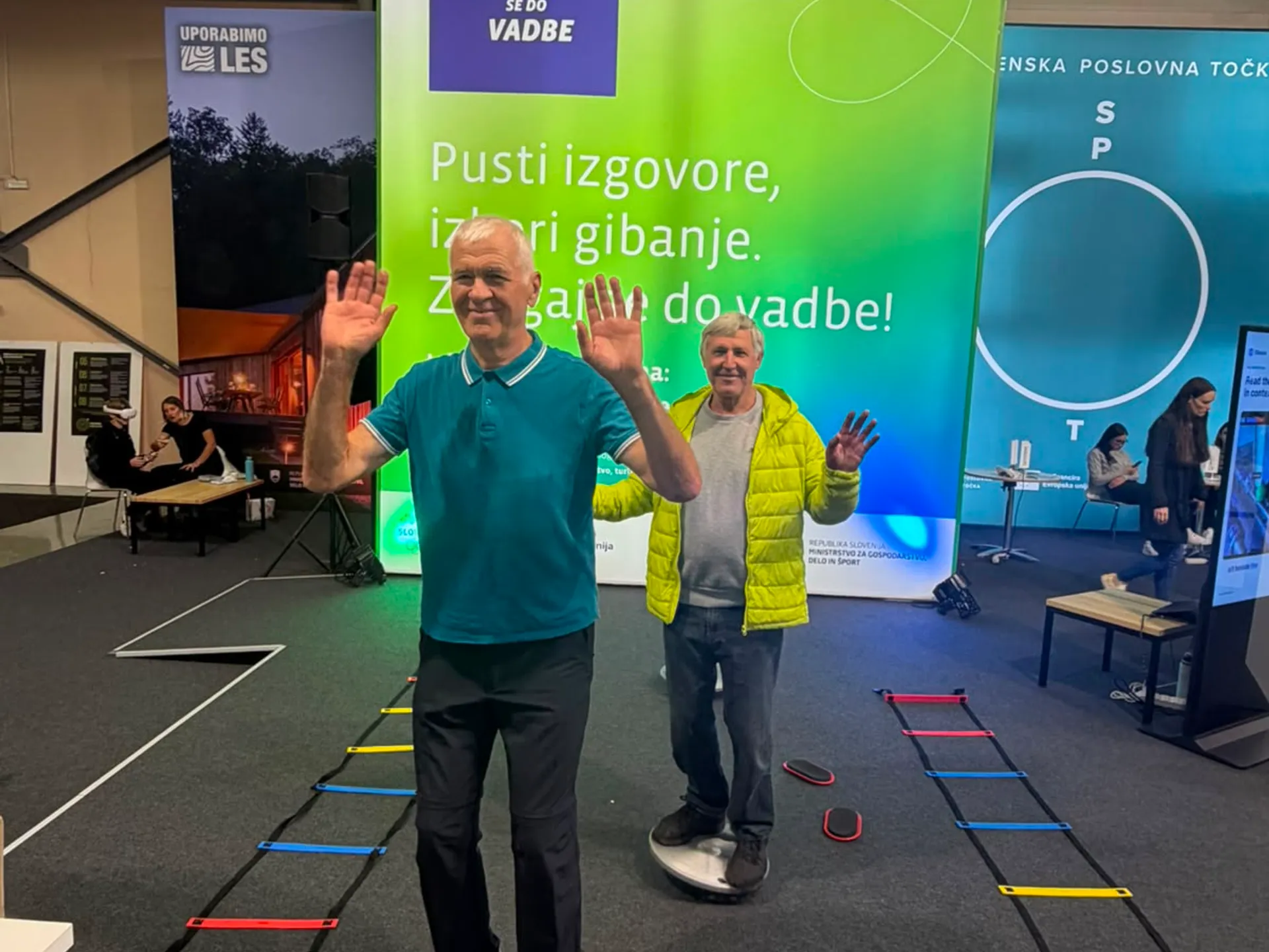Dva moška stojita na ravnotežnih ploščah, eden nosi modro majico, drugi svetlo rumeno jakno, ob njiju so različni športni pripomočki.