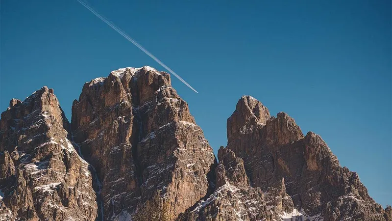 Visoki gorski vrhovi z nekaj snega na vrhu in letalska sled na modrem nebu.