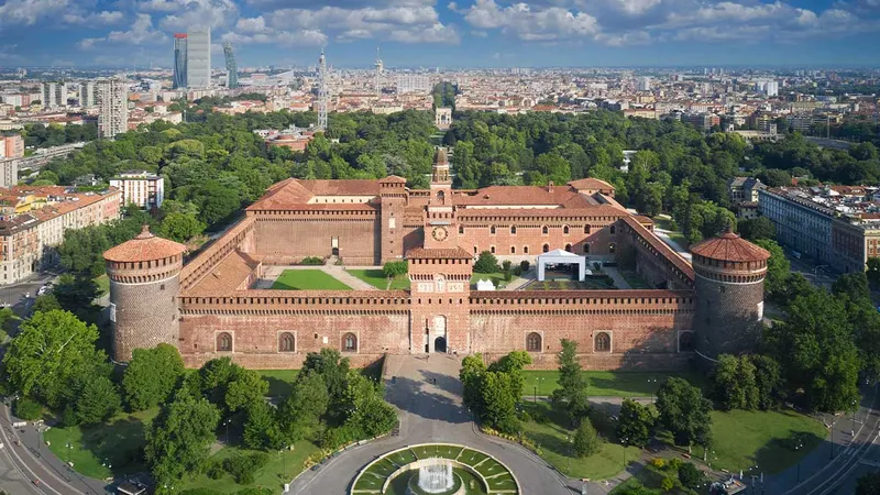 Grad Sforzesco z rdečo opeko in zelenimi površinami, obdan z drevesi in mestno arhitekturo v ozadju.