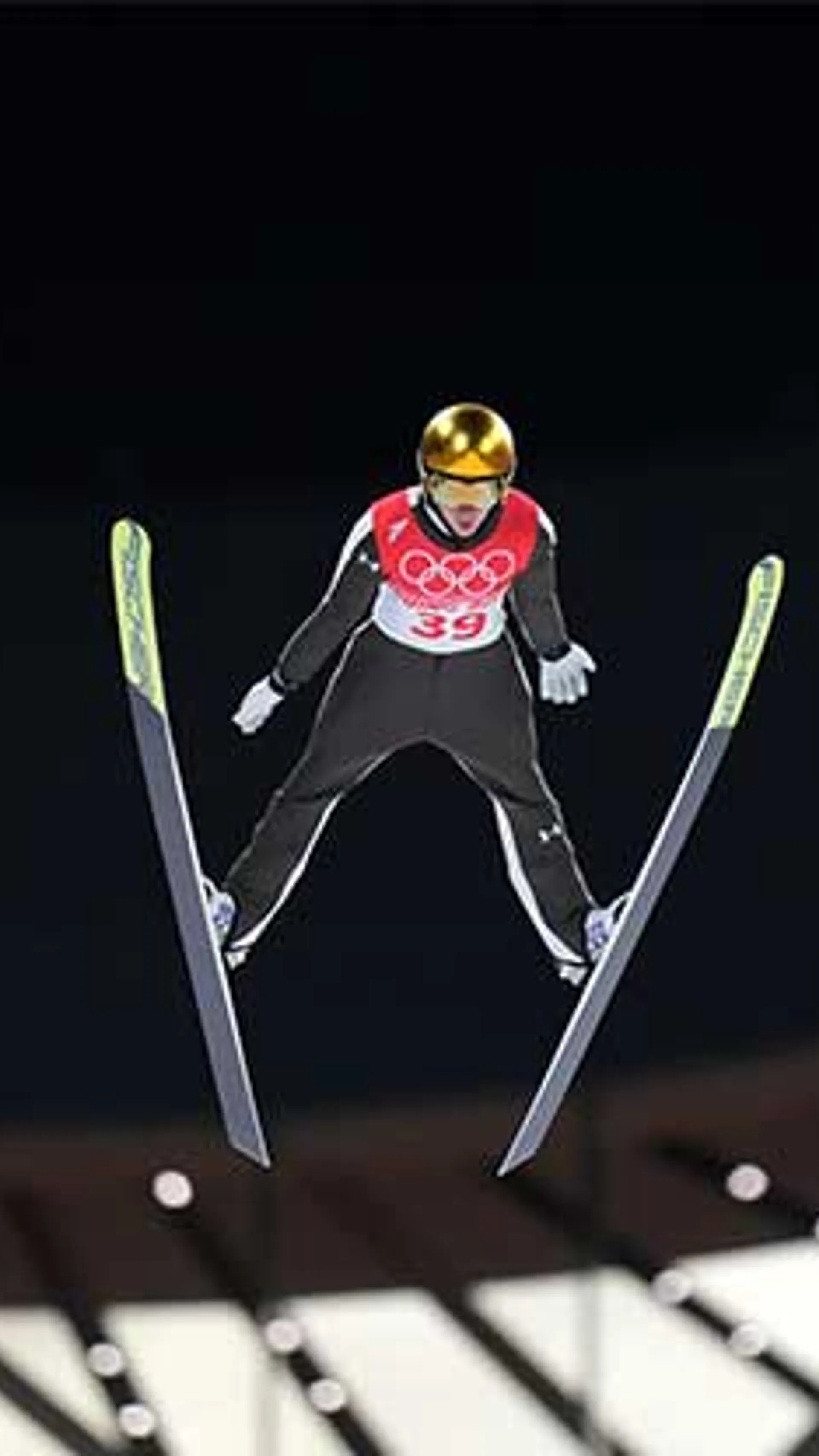 Smučar v zlatem čeladi, oblečen v črno-rdečo obleko, izvaja skok s smučmi, številka 39 na prsih.