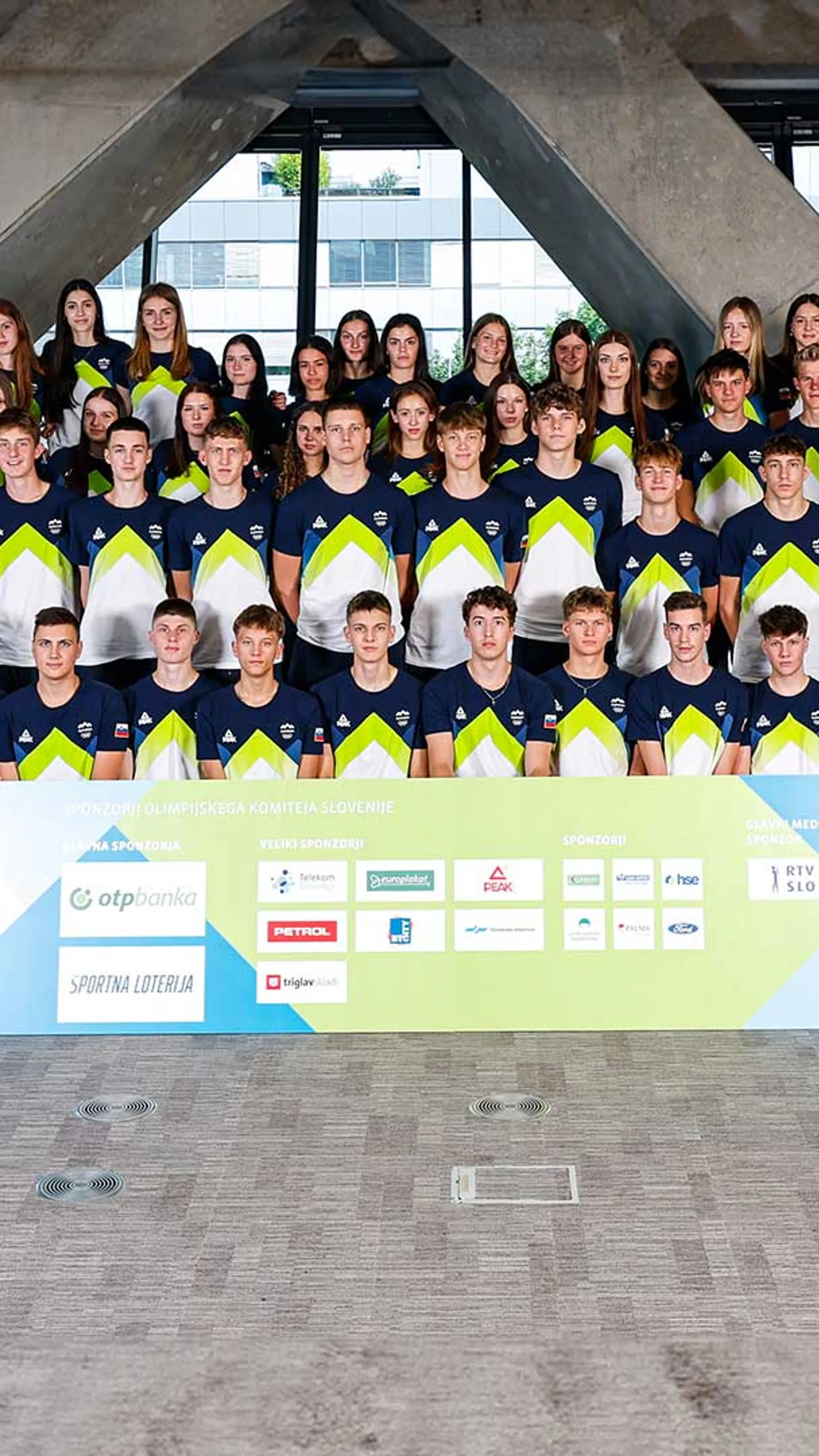 Skupinska fotografija športnikov v modrih in belih majicah z zelenimi detajli ter maskoto v oranžni obleki.