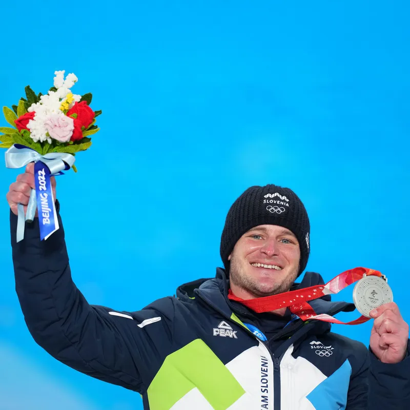 Tim Mastnak z nasmehom pozira s srebrno medaljo in šopkom ob ozadju z napisom Beijing 2022, kot simbol uspeha v deskanju na snegu v disciplini paralelni veleslalom