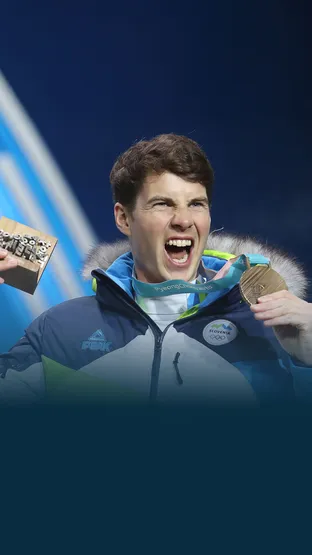 Žan Košir, športnik v slovenski zimski reprezentančni jakni navdušeno dviguje medaljo in leseno priznanje na svečani podelitvi.