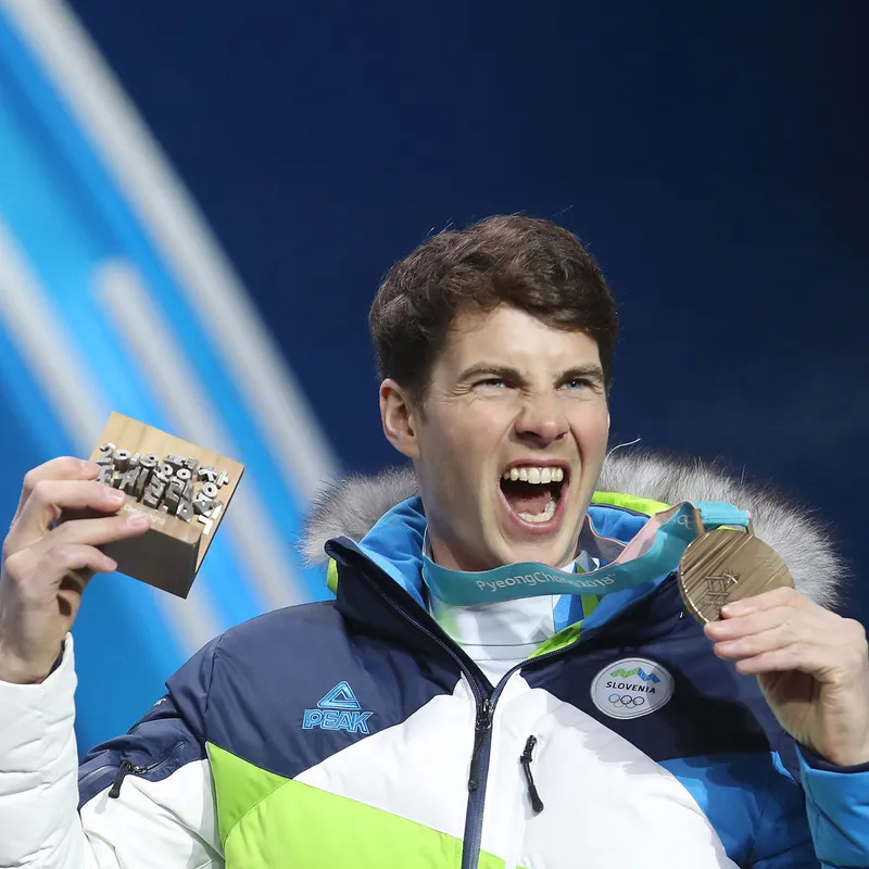 Žan Košir, športnik v slovenski zimski reprezentančni jakni navdušeno dviguje medaljo in leseno priznanje na svečani podelitvi.