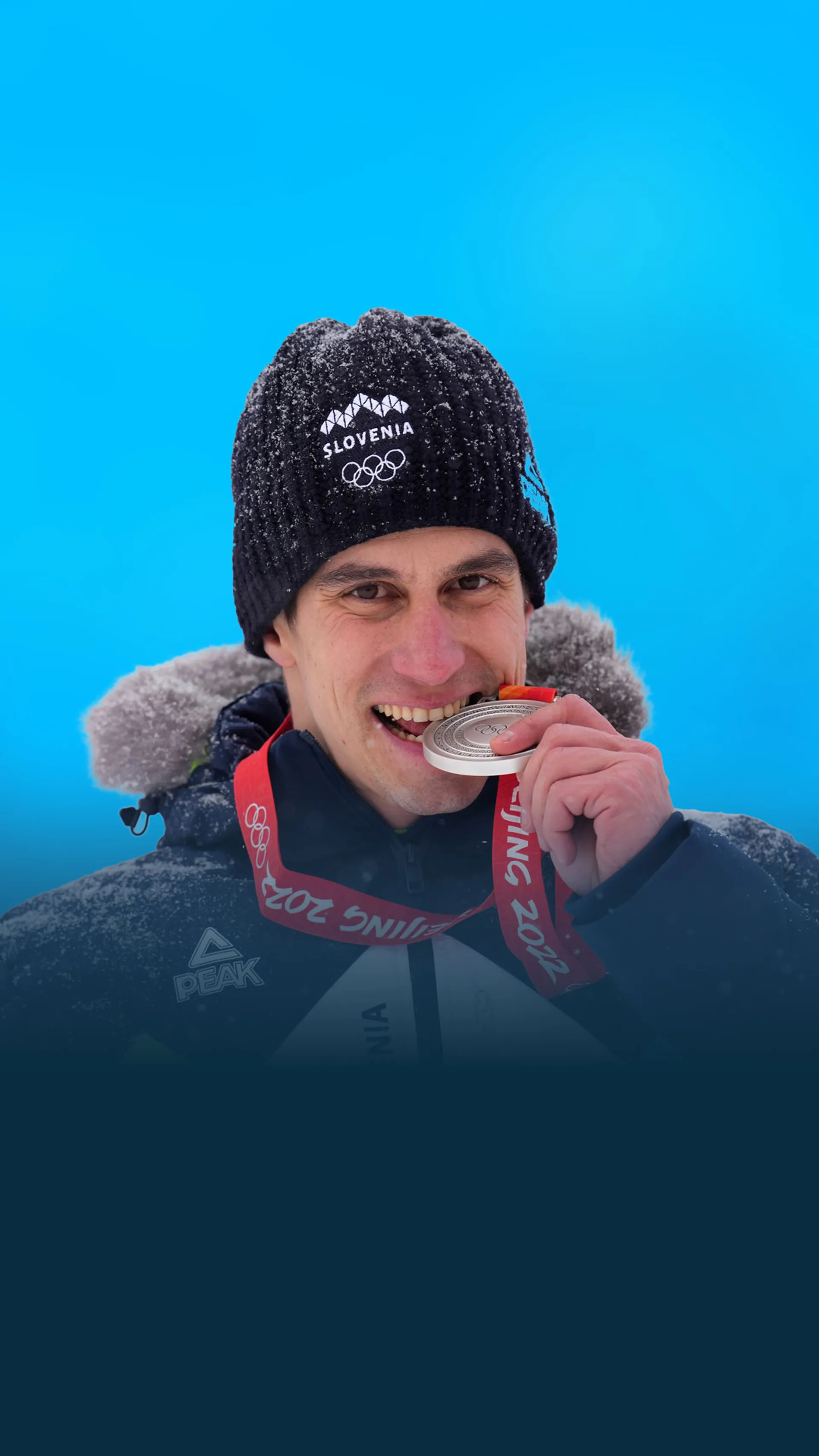 Žan Kranjec v slovenski zimski reprezentančni bundi stoji pred modrim ozadjem in ugrizne v srebrno medaljo s podelitve Beijing 2022.